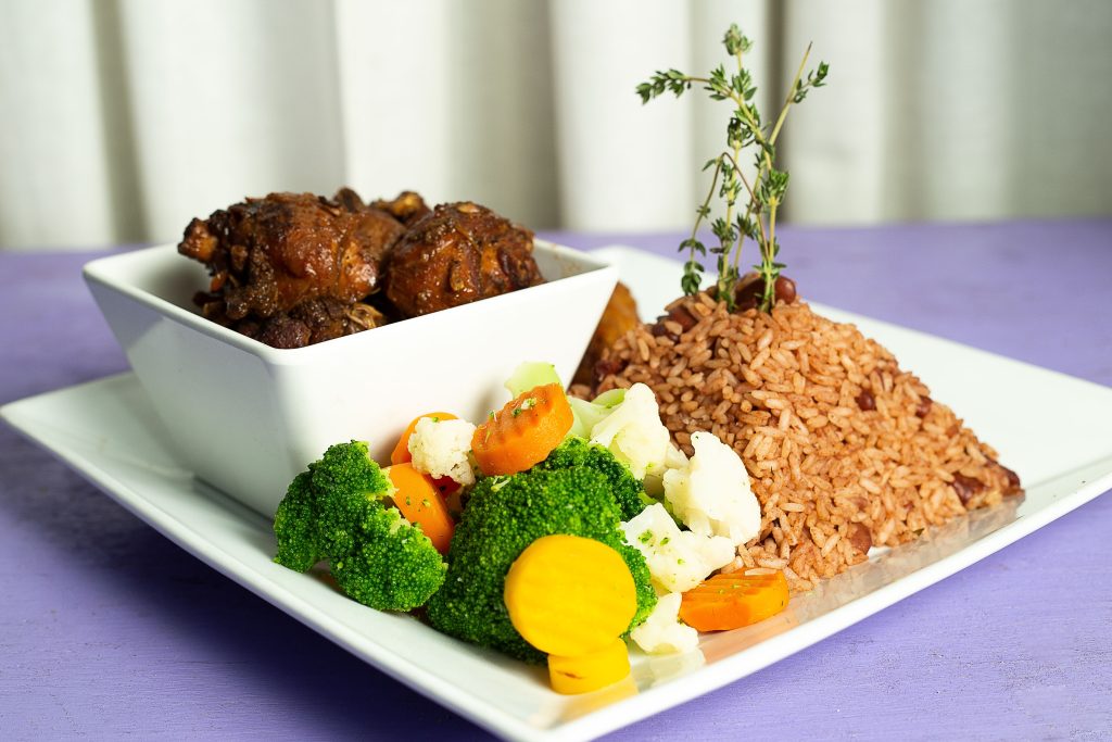 A plate with jollof rice, mixed vegetables, and a side of stewed meat, garnished with fresh herbs, on a purple table at one of the great food spots in Florida.