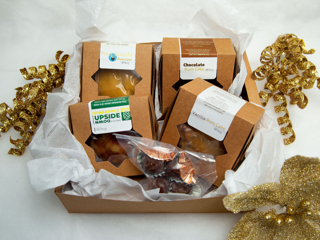 Assorted rum cakes in labeled boxes, including chocolate, vanilla, and pineapple flavors, displayed with shiny gold ribbons on a white background for the holiday season.