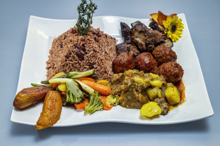 A plate with seasoned rice, roasted meat, meatballs, stewed vegetables, plantains, and a garnish of flowers and herbs celebrating Jamaican Independence.