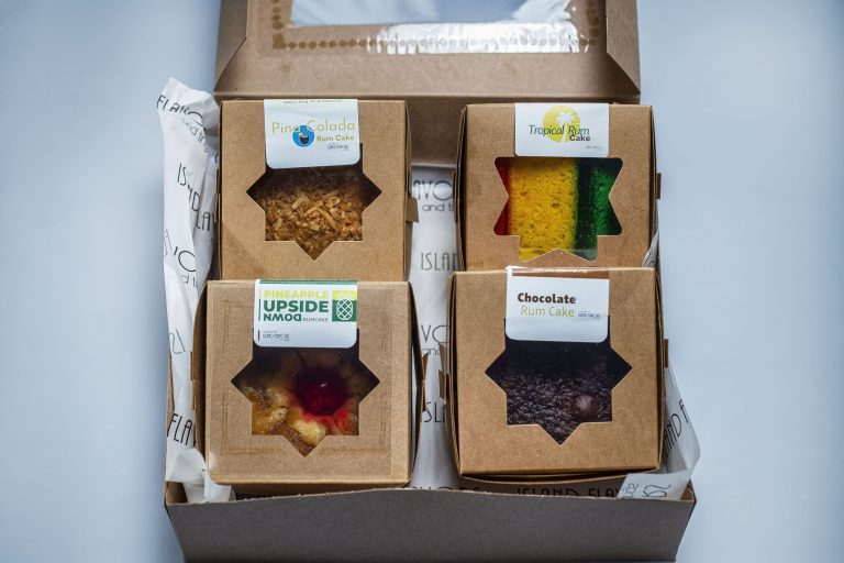 Four assorted cakes in a box labeled pina colada, tropical rum, pineapple upside down, and Jamaican Independence chocolate rum cake, viewed from above.