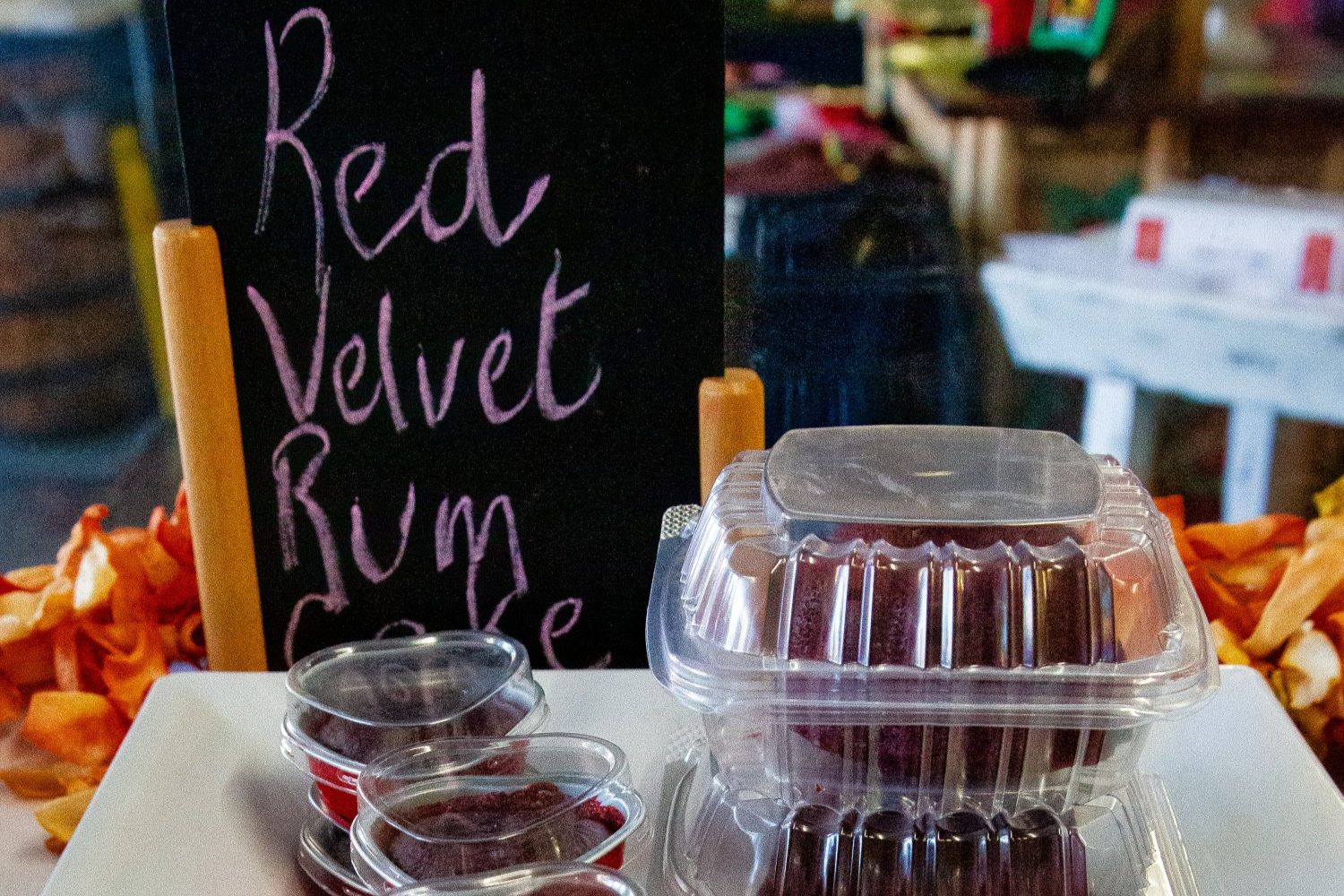 A table display featuring a sign that reads "Red Velvet Rumble" next to a container of red velvet rum cakes and small cups of red sauce.