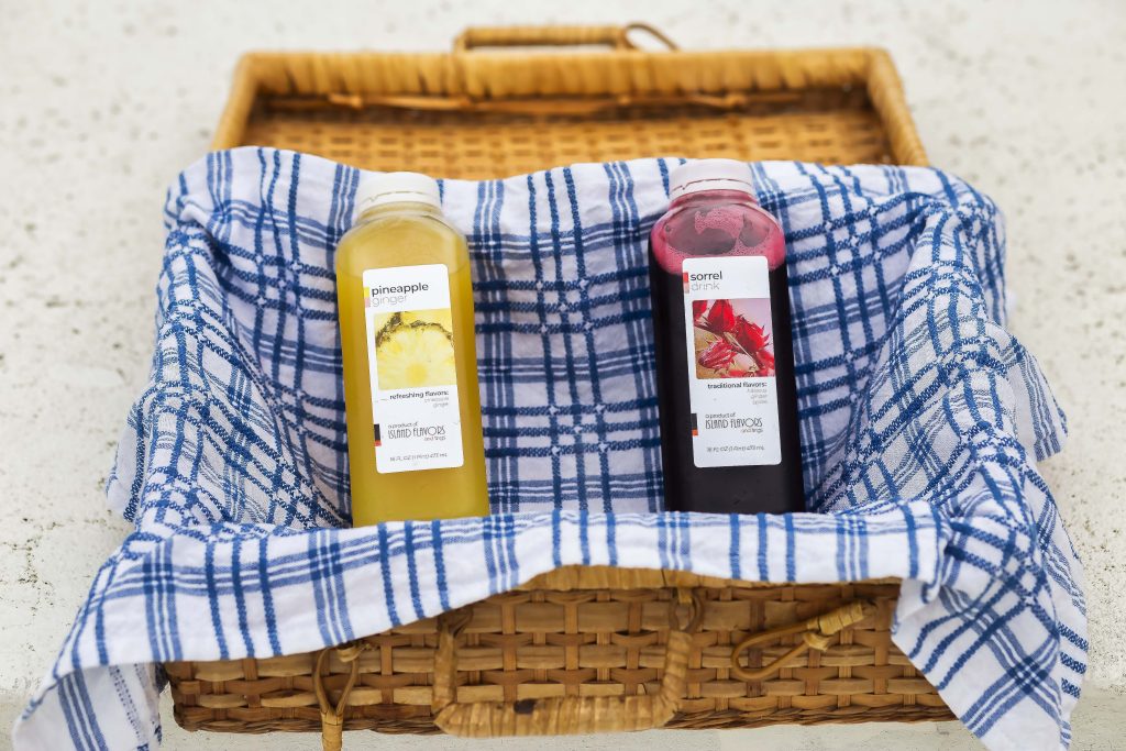 A wicker basket with a blue and white cloth contains two bottles of juice: one yellow labeled "pineapple" and one red labeled "sorrel." Perfect for a Summer Refreshments