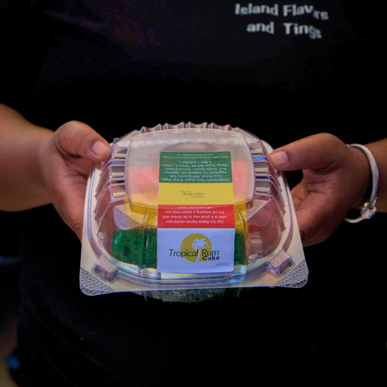 Person holding a clear plastic container of Tropical Rum Cake with a label on top displaying the name and description of the product, celebrating Jamaican Independence.