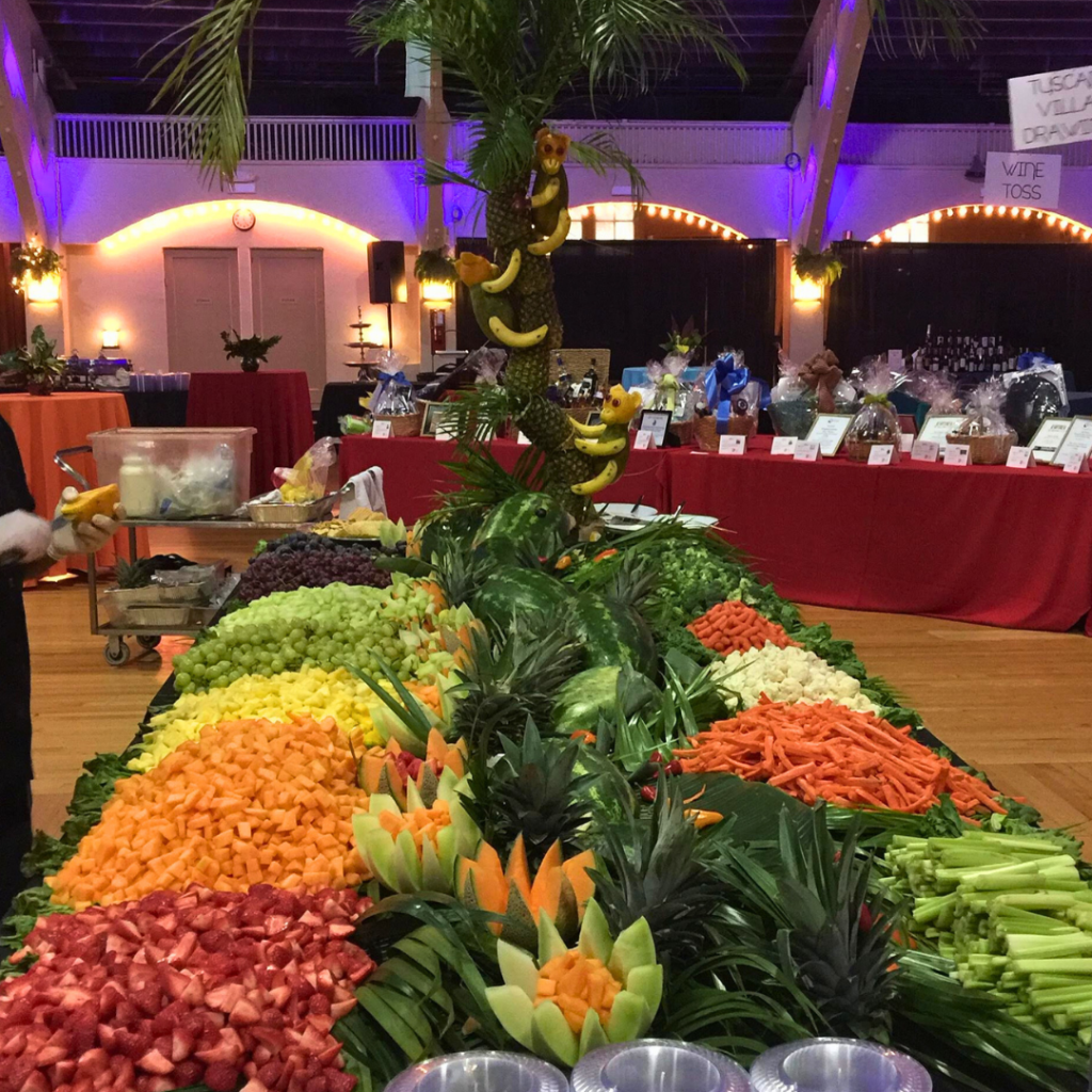 A large display of assorted chopped fruits and vegetables, perfect for corporate catering during the holiday season, is arranged on a long table indoors with a decorative tree centerpiece. Fruits include melon, grapes, and strawberries; vegetables include carrots and celery—a hit among busy professionals.