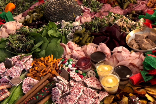 A table laden with a variety of foods, perfect for holiday catering, includes sliced meats, greens, olives, pickles, candies, pretzels, breadsticks, chocolates, and three small candles in the foreground. This setup offers menu ideas that will surely wow your guests.