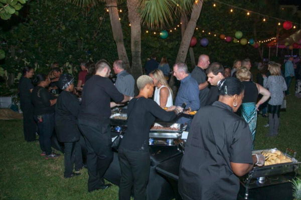 People gather around a buffet table at an outdoor event under string lights and trees with colorful lanterns hanging. Several individuals serve food while others wait in line or chat, inspired by holiday catering menu ideas designed to wow your guests.