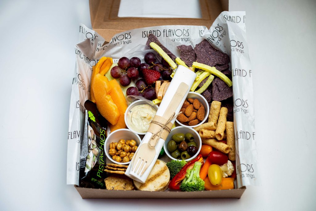 A cardboard box contains assorted snacks—perfect for foodies—including sliced fruits, grapes, vegetables, nuts, crackers, chips, dip, and utensils on branded paper; an ideal pick for any holiday gift guide.