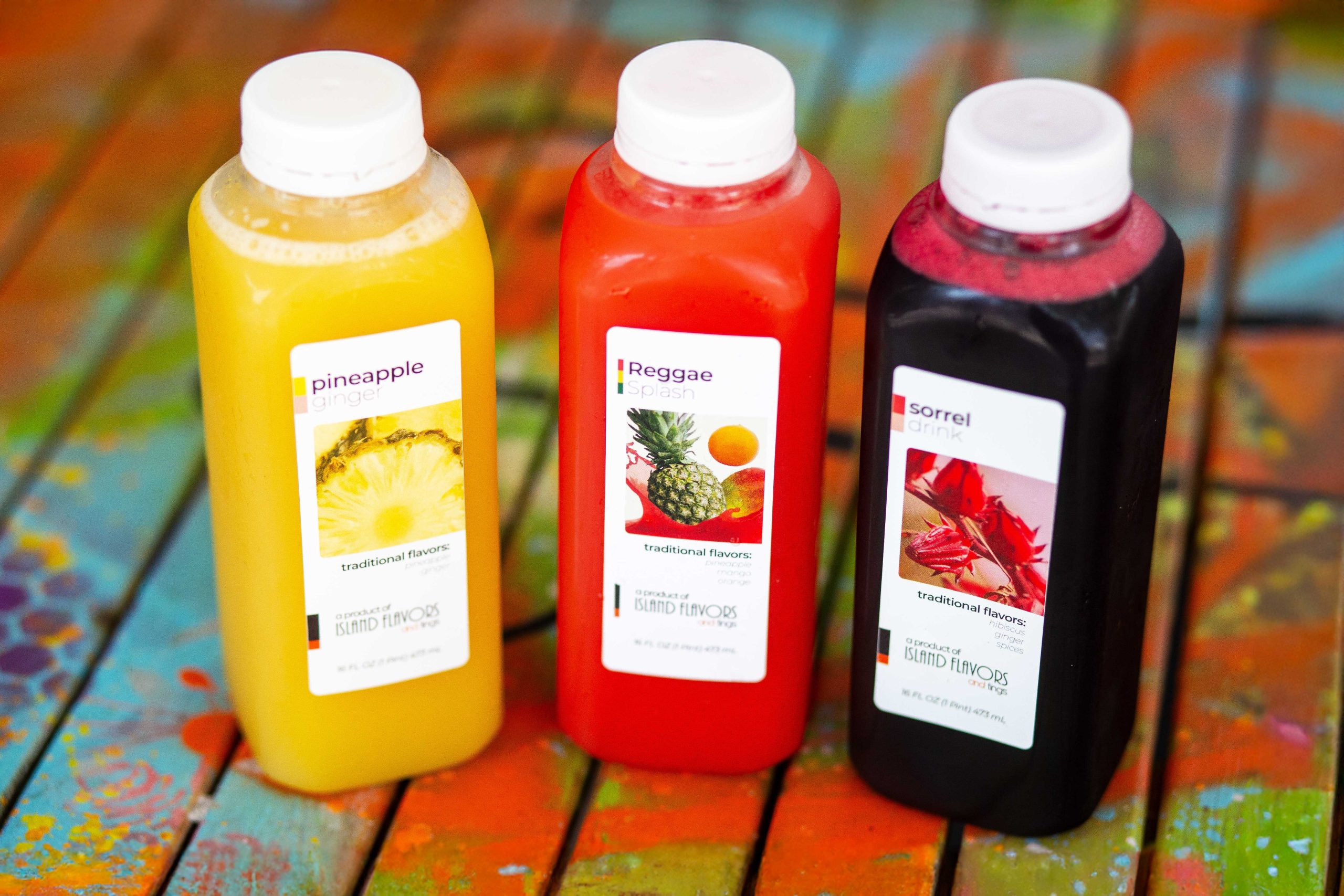 Three plastic bottles of juice labeled Pineapple, Reggae, and Sorrel sit on a colorful wooden surface at a Jamaican restaurant. Each label features fruit images and the phrase "traditional flavors.