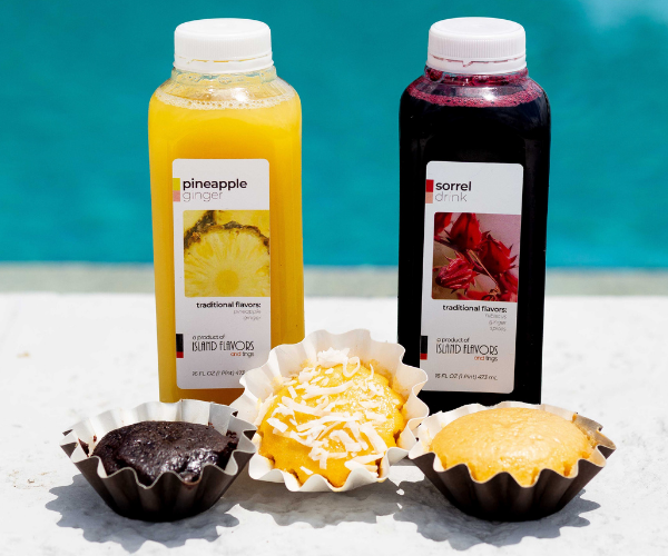 Two bottled drinks, one labeled "pineapple ginger" and the other "sorrel drink," are placed behind three cupcakes of different flavors with toppings, set on a white surface near a pool.
