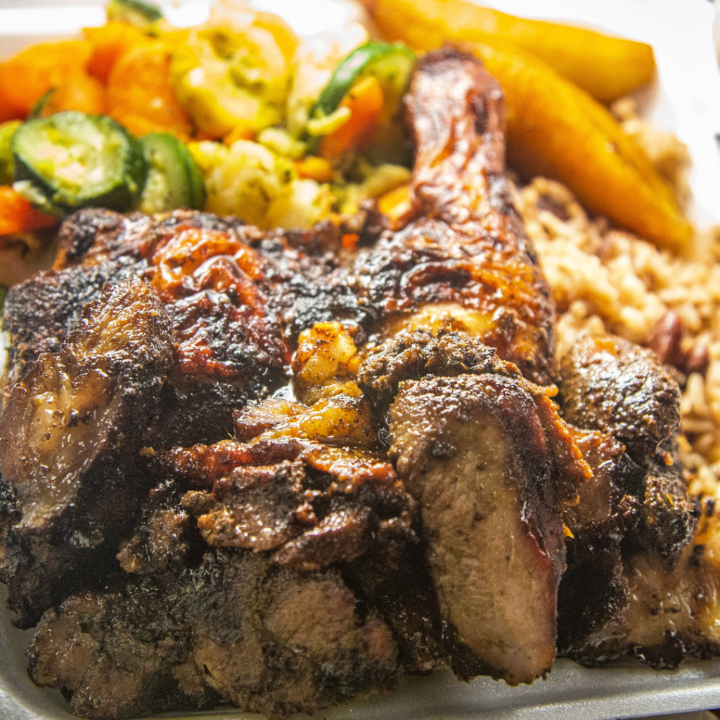 A close-up of a takeout container with grilled jerk chicken, rice and peas, sautéed vegetables, and fried plantains—perfect for homestyle meals or sharing as family meals.