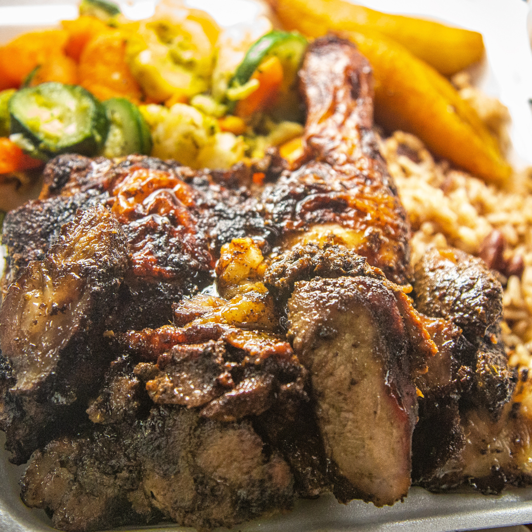 A close-up of a takeout container with grilled jerk chicken, rice and peas, sautéed vegetables, and fried plantains—perfect for homestyle meals or sharing as family meals.